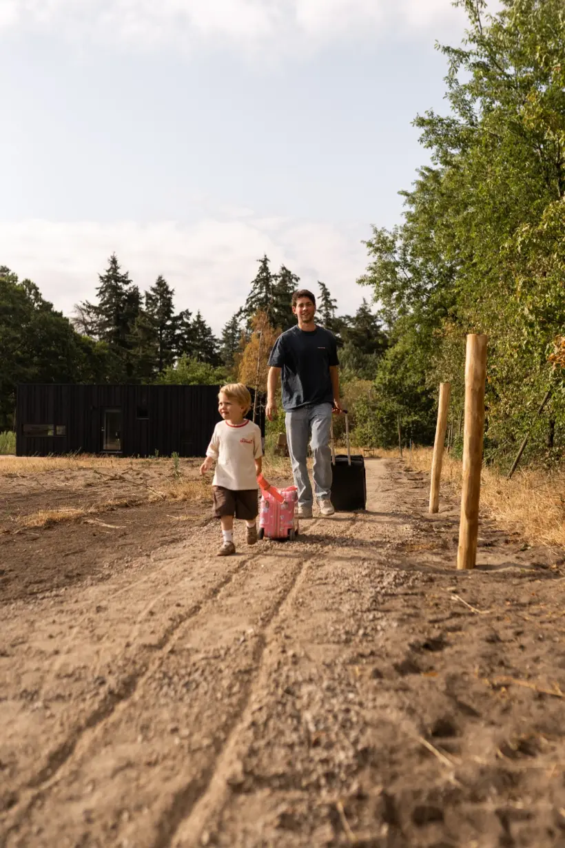 Een volwassene en een klein kind lopen met hun koffers over het zandpad bij de lodges