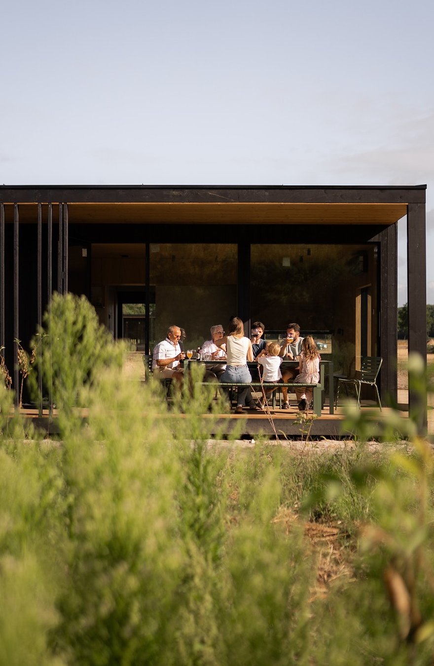 Eco Lodge De Adem met gasten aan tafel op het terras, gezien vanaf de wilde beplanting ervoor