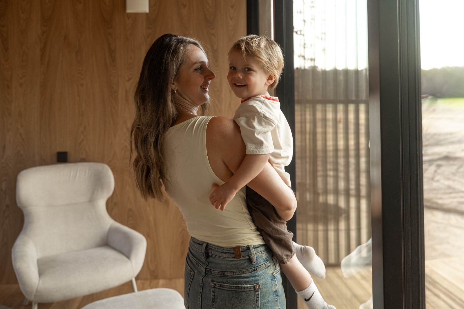 Vrouw met een peuter op de arm bij de grote raampartij van de woonruimte